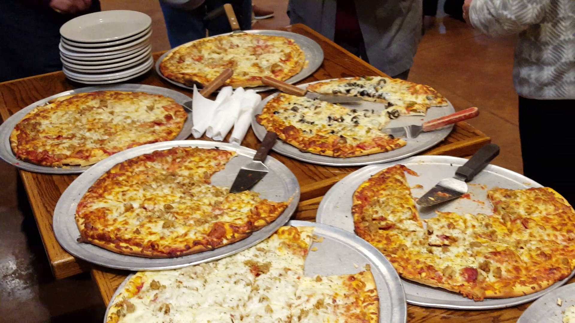 2 tables with 6 different pizzas, serving utensils, plates, and silverware.